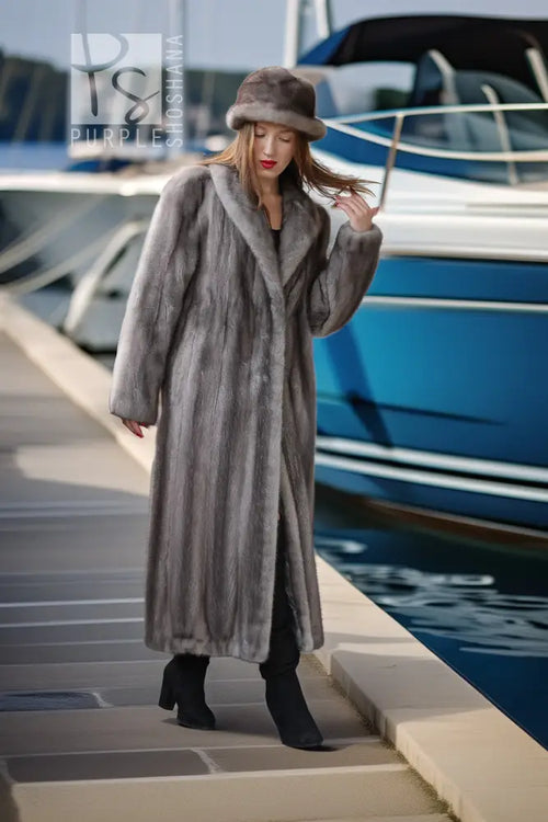 A woman wearing a long gray faux fur coat, matching gray fur hat, and black boots walks along a concrete dock next to a blue and white yacht.