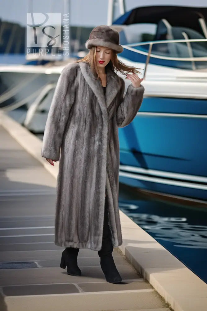 A woman wearing a long gray faux fur coat, matching gray fur hat, and black boots walks along a concrete dock next to a blue and white yacht.