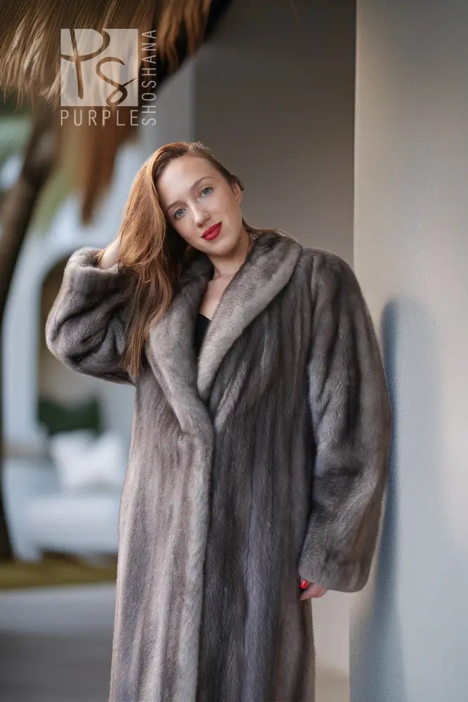 A woman wearing a luxurious, long gray fur coat with a wide collar, posing in a softly lit indoor setting with a thatched roof visible in the background.