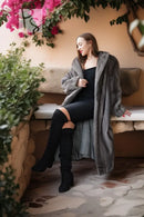 A woman sits on a stone bench wearing a long gray faux fur coat over a black dress and black over-the-knee boots, with pink flowers and green plants in the background.