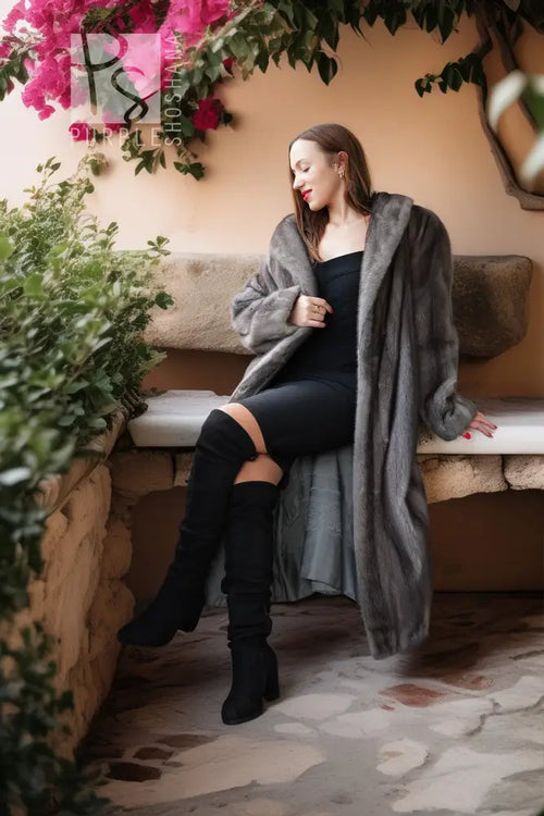 A woman sits on a stone bench wearing a long gray faux fur coat over a black dress and black over-the-knee boots, with pink flowers and green plants in the background.