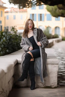A woman sits on a stone ledge outdoors wearing a long gray faux-fur coat over a black dress and black over-the-knee boots, with a blurred building and water in the background.
