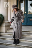 A woman wearing a long, luxurious gray fur coat, matching gray fur hat, and black boots stands on stone steps in front of a building with a blue door.