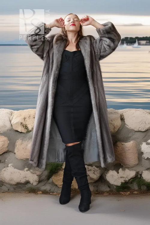 A woman wearing a long gray faux fur coat over a black dress and black knee-high boots, posing outdoors in front of a stone wall with water and a cloudy sky in the background.