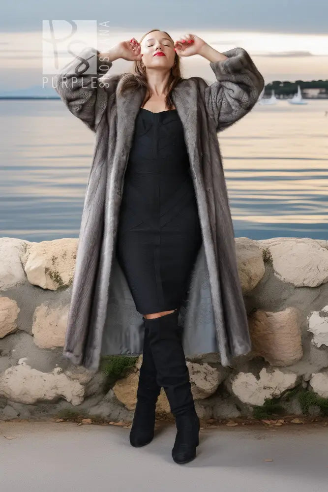 A woman wearing a long gray faux fur coat over a black dress and black knee-high boots, posing outdoors in front of a stone wall with water and a cloudy sky in the background.