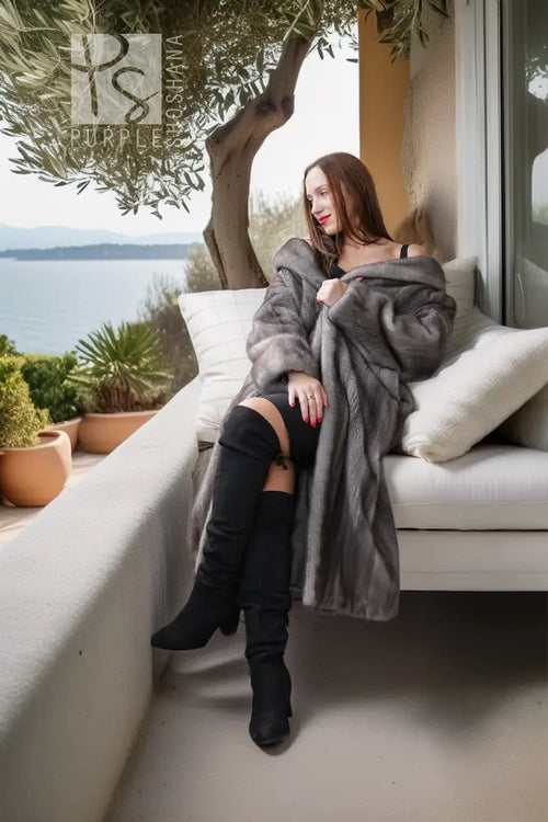 A woman sits on a white outdoor sofa on a balcony overlooking water, wearing a long gray fur coat and black over-the-knee boots.