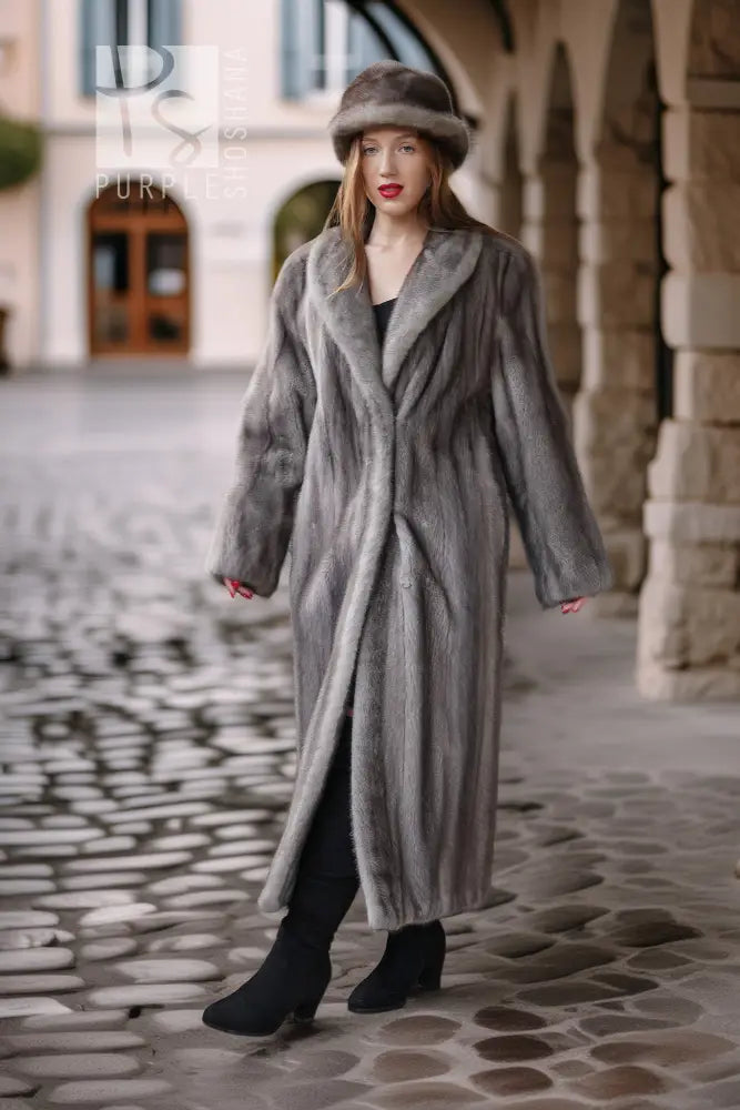 A woman wearing a long gray fur coat and matching fur hat stands on a cobblestone street in front of a stone archway building.