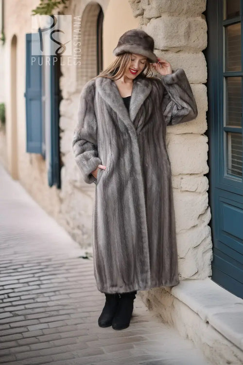 A woman wearing a long gray fur coat and matching fur hat stands on a cobblestone street beside a stone building with blue shutters.