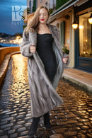 A woman walking on a cobblestone street at dusk, wearing a long gray fur coat over a black dress and black boots.