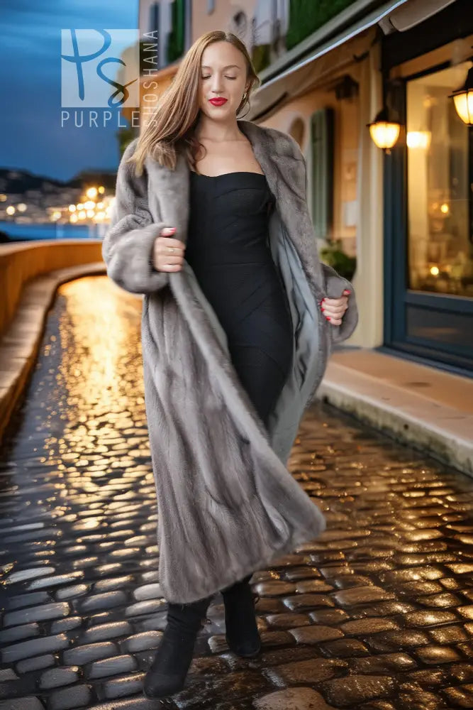A woman walking on a cobblestone street at dusk, wearing a long gray fur coat over a black dress and black boots.