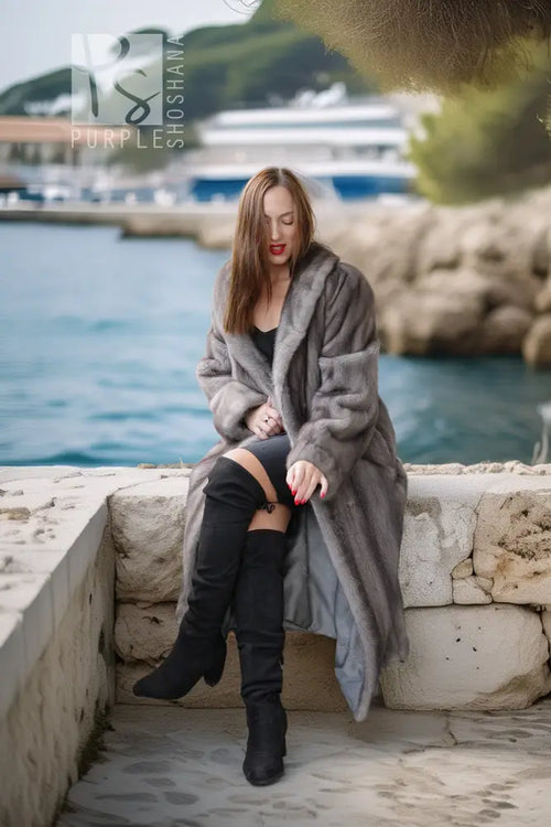 A woman sits on a stone ledge by the water wearing a long gray faux fur coat, black thigh-high boots, and a black top. The background shows a coastal marina with boats and green trees.