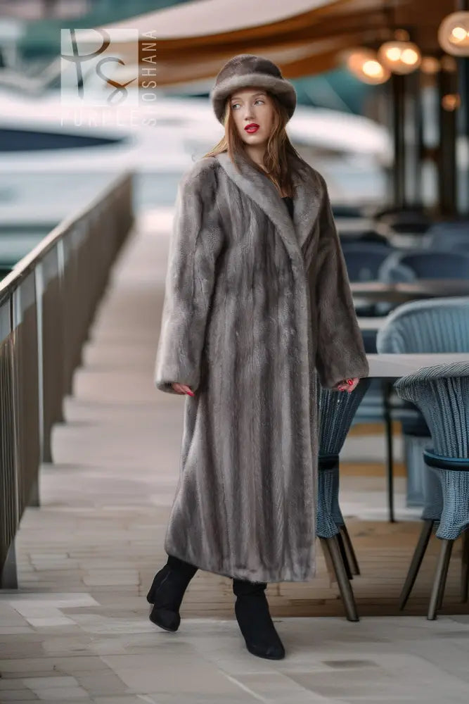 A woman models a long, luxurious gray fur coat and matching fur hat on an outdoor deck next to water, with a blurred background of boats and patio seating.