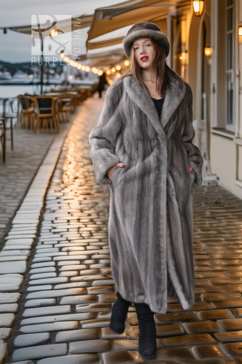 A woman wearing a long gray fur coat, matching fur hat, and black boots stands on a cobblestone street at dusk, with warm lights and outdoor seating in the background.