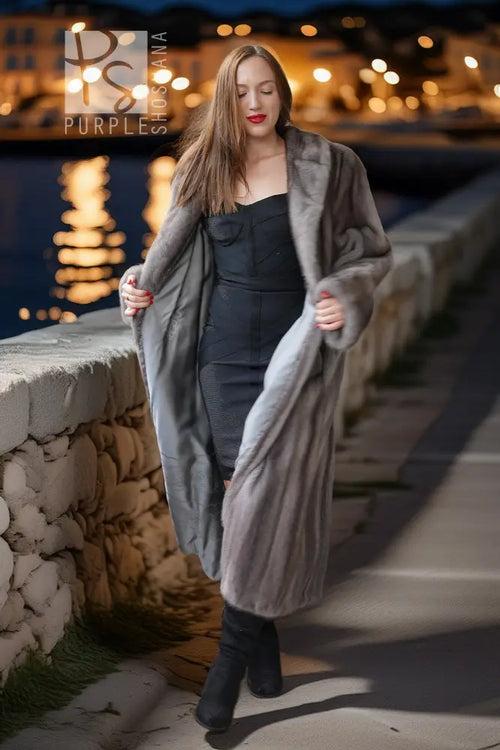 A woman wearing a long, plush gray fur coat over a black strapless dress and black boots, standing on a stone walkway at night with warm bokeh lights and water in the background.