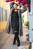 A woman stands in an outdoor alley wearing a long gray faux fur coat over a black dress and black over-the-knee boots, with bright pink flowers hanging overhead.