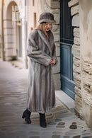 A woman wearing a long gray fur coat, matching fur hat, and black ankle boots stands on a cobblestone street in front of a stone building with a blue door.