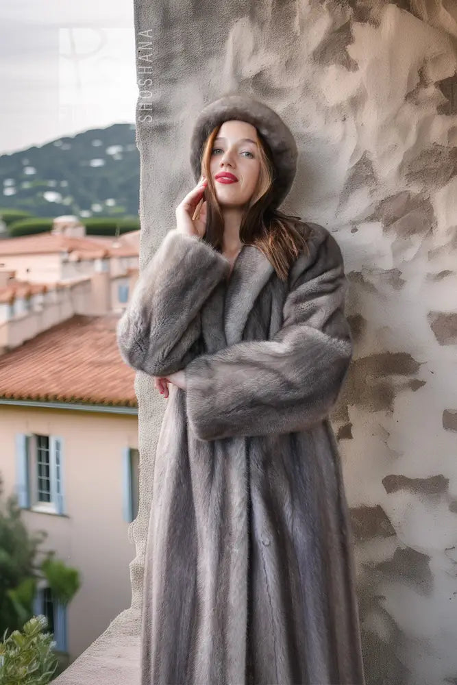 A woman wearing a luxurious gray fur coat and matching fur hat leans against a textured stone wall, with a scenic Mediterranean village in the background.