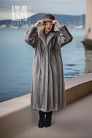 A woman wearing a long gray fur coat, matching fur hat, and black boots stands on a waterfront promenade with water and distant hills in the background.
