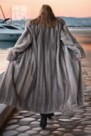 A woman viewed from behind wearing a long, voluminous gray fur coat while walking on a stone pier by the water at sunset.