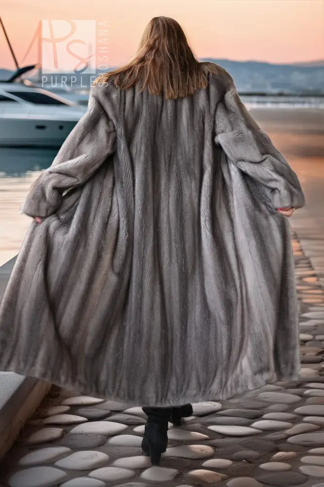 A woman viewed from behind wearing a long, voluminous gray fur coat while walking on a stone pier by the water at sunset.