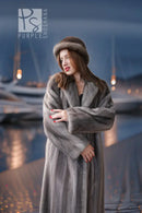A woman wearing a luxurious gray fur coat and matching fur hat stands on a waterfront promenade at dusk, with boats and marina lights blurred in the background.