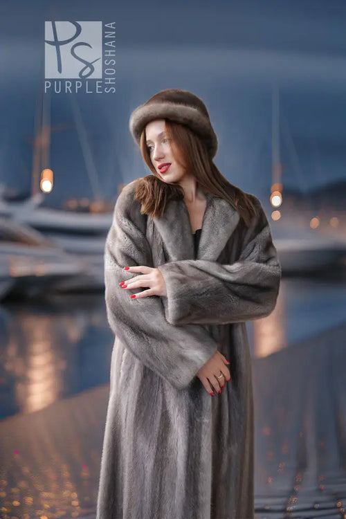 A woman wearing a luxurious gray fur coat and matching fur hat stands on a waterfront promenade at dusk, with boats and marina lights blurred in the background.