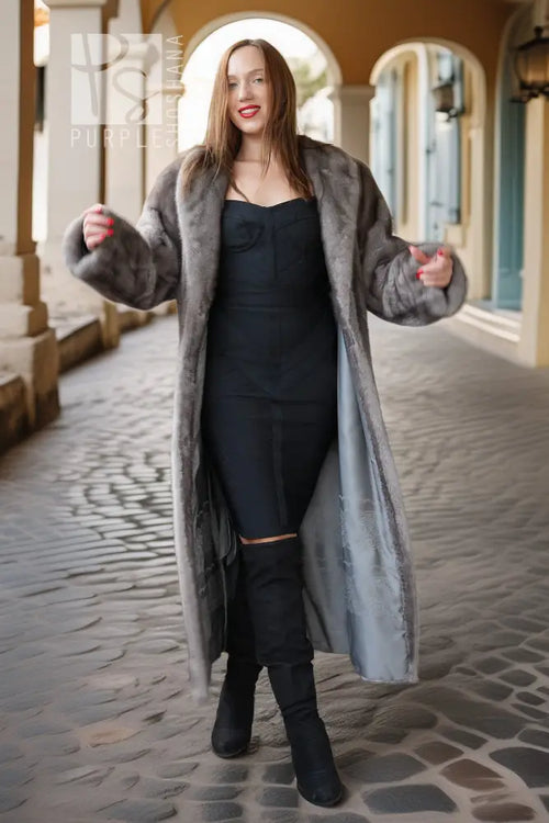 A woman stands in a cobblestone walkway between arches, wearing a long gray faux-fur coat over a black dress and black over-the-knee boots.