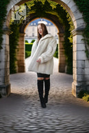 A woman wearing a fluffy cream faux-fur coat, black shorts, and black over-the-knee boots walks through a stone archway covered in green ivy toward a grand building in the background.