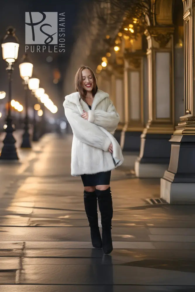 A woman wearing a white faux fur coat, black mini skirt, and black over-the-knee boots walks down a city street at night with warm streetlights and building columns in the background.