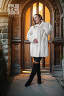A woman wearing a voluminous white faux fur coat, black distressed leggings, and black over-the-knee boots stands in front of an ornate stone and wood arched doorway.
