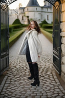 A woman stands in a stone courtyard framed by open black iron gates, wearing a voluminous off-white faux fur coat over a dark dress and black over-the-knee boots. A chateau and manicured hedges are visible in the background.
