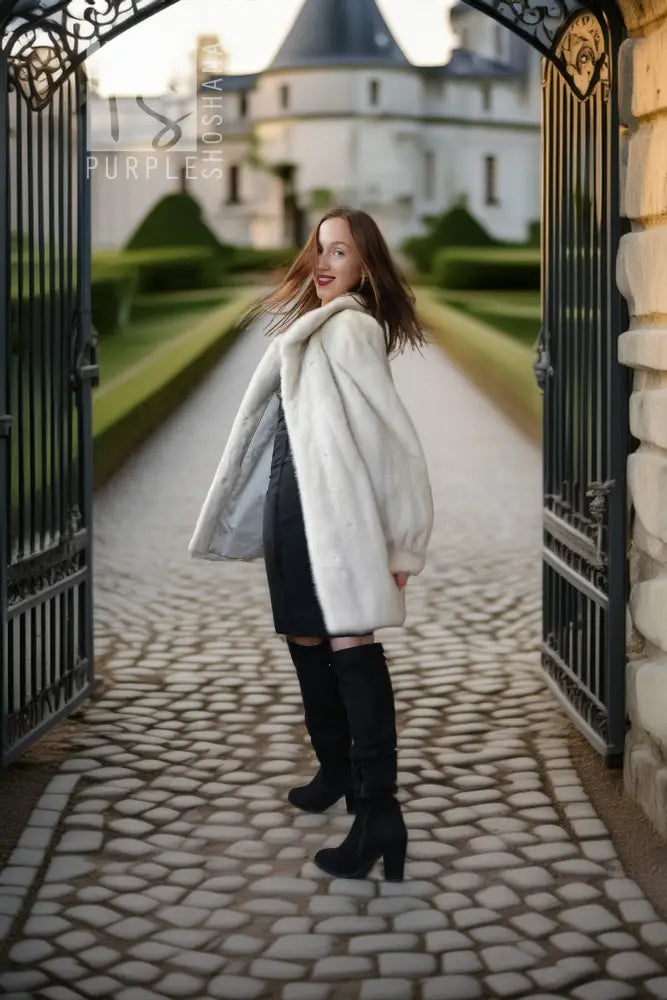 A woman stands in a stone courtyard framed by open black iron gates, wearing a voluminous off-white faux fur coat over a dark dress and black over-the-knee boots. A chateau and manicured hedges are visible in the background.