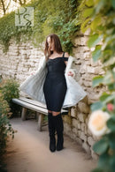 A woman stands outdoors on a stone path beside a stone wall with greenery, wearing a fitted black dress, a light gray coat, and black knee-high boots.