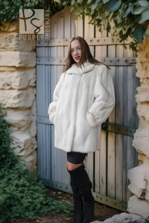 A woman wearing a voluminous white faux fur coat, black leggings, and black over-the-knee boots stands in front of a weathered gray wooden door framed by stone walls and green ivy.