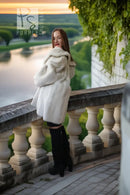 A woman wearing a voluminous white fur coat and black over-the-knee boots stands on a stone terrace overlooking a scenic lake and green landscape at sunset.