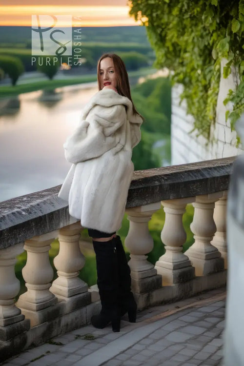 A woman wearing a voluminous white fur coat and black over-the-knee boots stands on a stone terrace overlooking a scenic lake and green landscape at sunset.