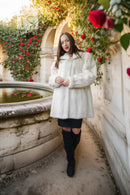 A woman wearing a voluminous white faux fur coat, black skirt, and black over-the-knee boots stands in a sunlit garden corridor with a stone fountain and red rose vines.