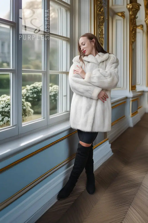 A woman stands by a large window in an ornate room, wearing a voluminous white faux fur coat, black leggings, and black over-the-knee boots.