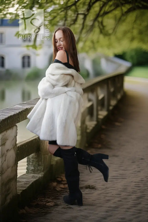 A woman posing on a stone bridge outdoors, wearing a voluminous white faux fur coat over a black outfit with black over-the-knee boots.