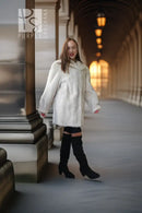 A woman wearing a voluminous white fur coat, black thigh-high cutout boots, and black knee-high socks walks through a covered outdoor corridor with warm hanging lights in the background.