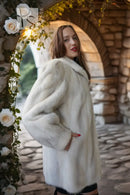 A woman wearing a voluminous white fur coat stands in a stone archway decorated with white roses and green vines.