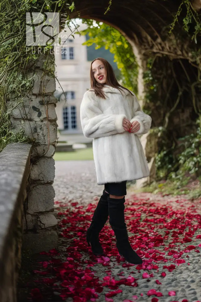 A woman wearing a white faux fur coat, black leggings, and black over-the-knee boots stands under a stone archway with red rose petals on the ground.