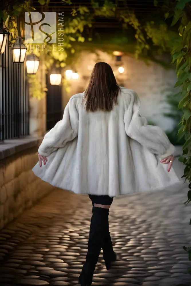 A woman viewed from behind wearing a voluminous white faux fur coat, black pants, and black boots, walking on a cobblestone path under a stone archway with hanging lanterns and green ivy.